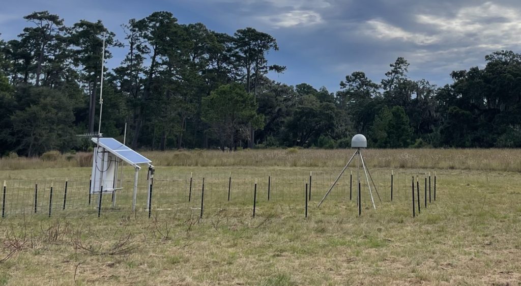 A CORS station with a solar panel and a GNSS receiver within a fence and field