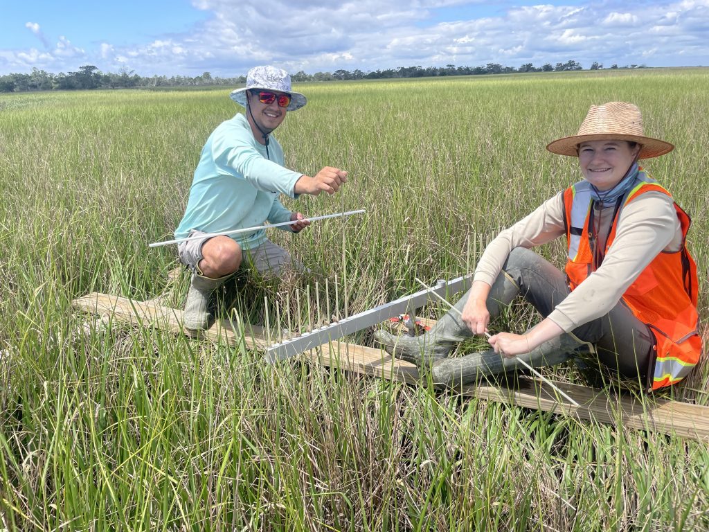 Two people smiling, sitting on a wooden platform in saltmarsh with scientific measuring equipment